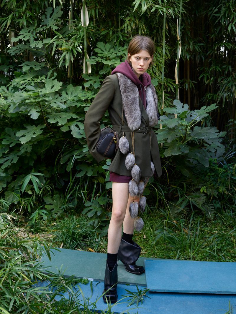 A fashionable adult female stands in a green outdoor setting, wearing an olive jacket, hooded top, black boots, and carrying a small shoulder bag.