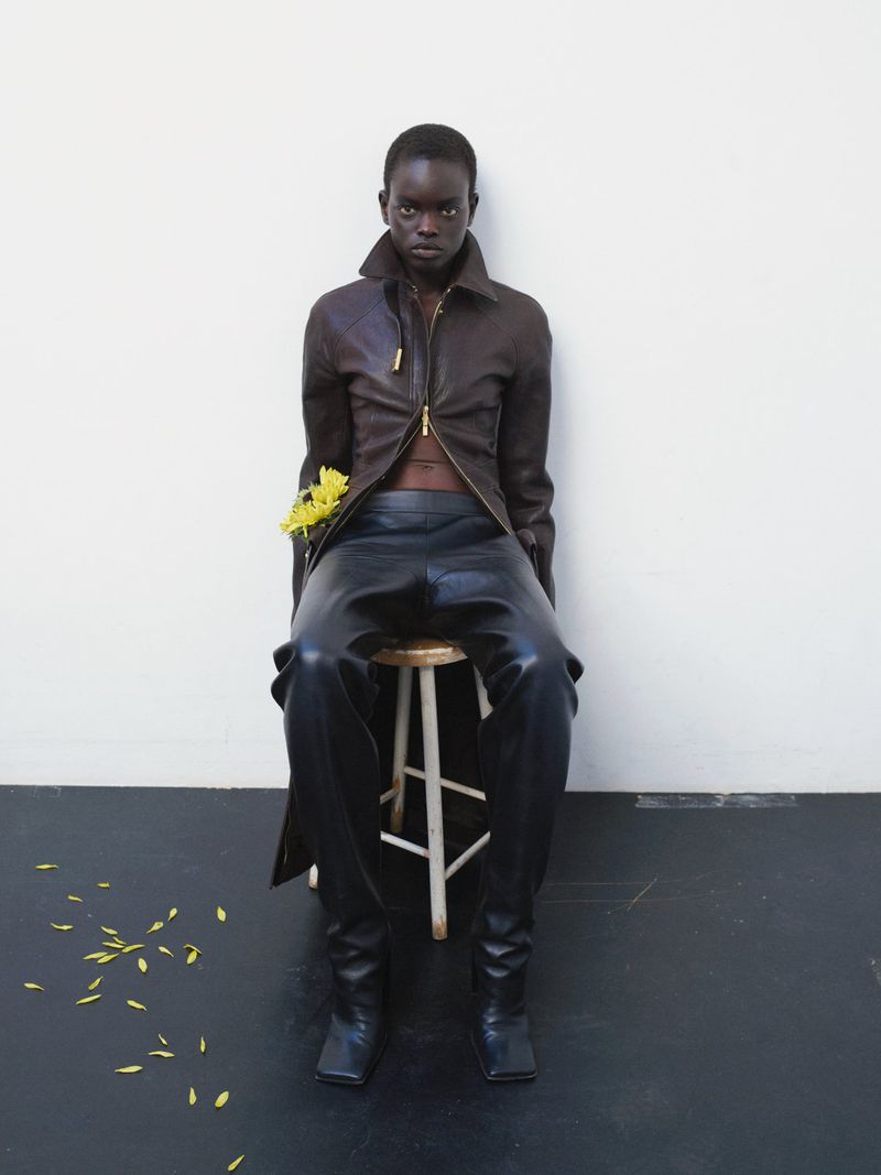 A woman with dark skin and a serious expression models a dark brown jacket and black leather pants while seated on a wooden stool.