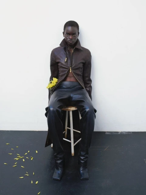 A woman with dark skin and a serious expression models a dark brown jacket and black leather pants while seated on a wooden stool.