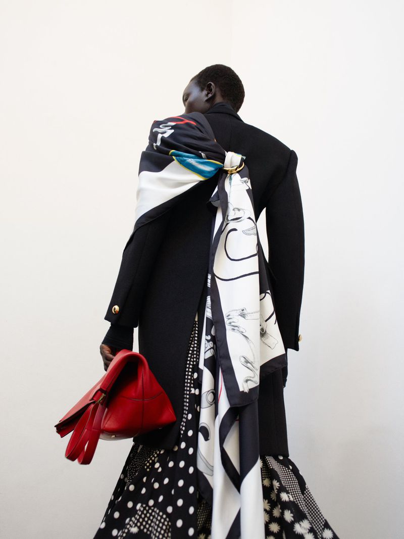 Woman from the back in a black blazer with a patterned scarf and skirt, holding a red bag in a minimalist white setting.