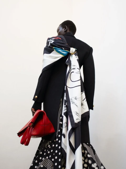 Woman from the back in a black blazer with a patterned scarf and skirt, holding a red bag in a minimalist white setting.