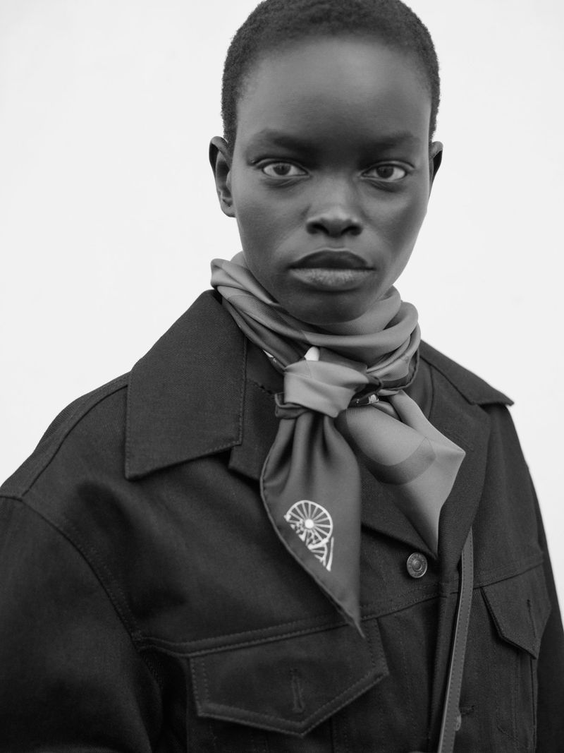 Black and white portrait of a woman wearing a dark collared jacket and a printed silk neck scarf against a pale overcast sky.
