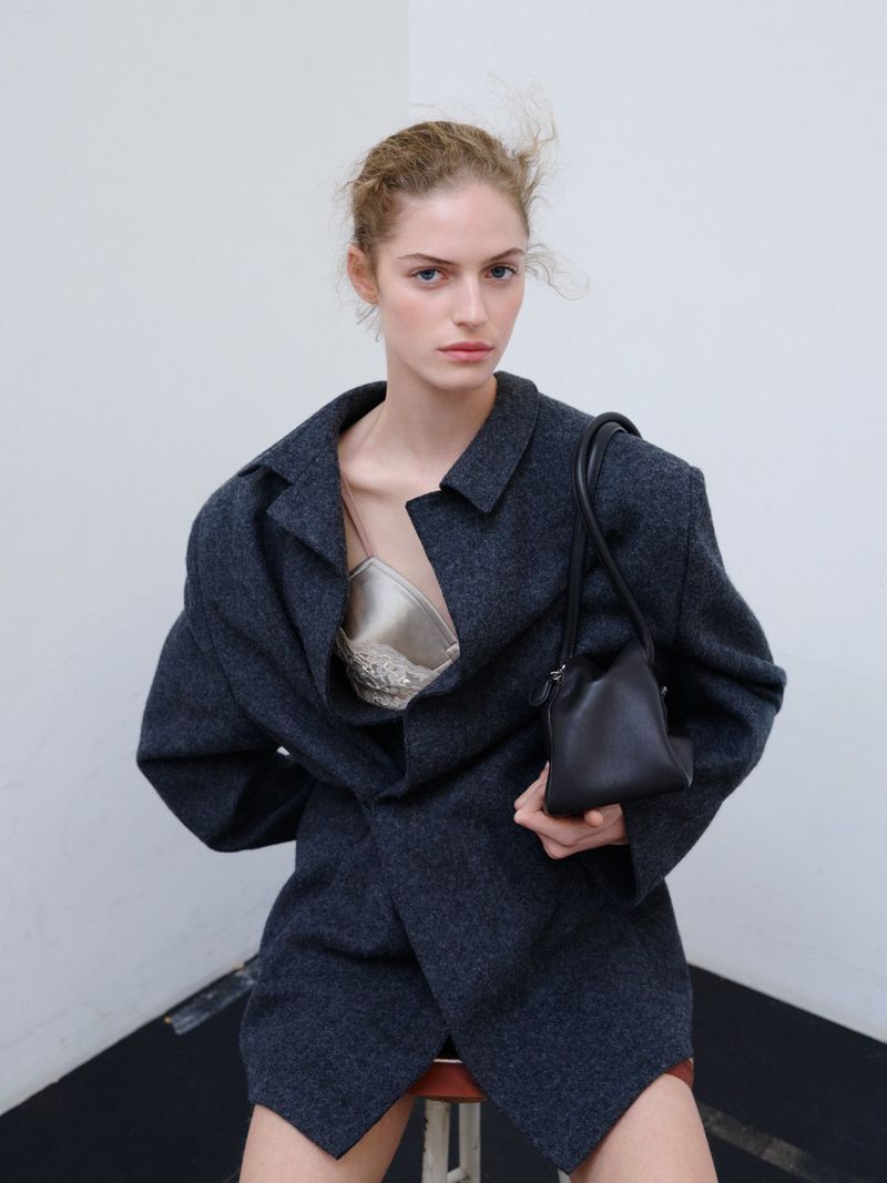 A female fashion model wearing a blazer and holding a small black handbag poses in a corner against a white wall in a studio.