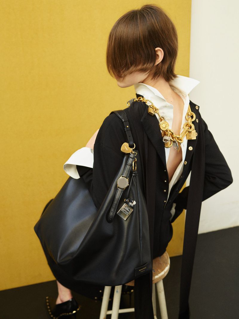 Model seated on a stool with an oversized black leather shoulder tote, open-back white collared shirt, and chunky gold chain necklace.