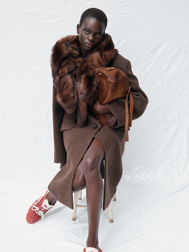 Portrait of a Black fashion model in a brown skirt suit with a fur collar and purse, seated against a white backdrop in a studio.