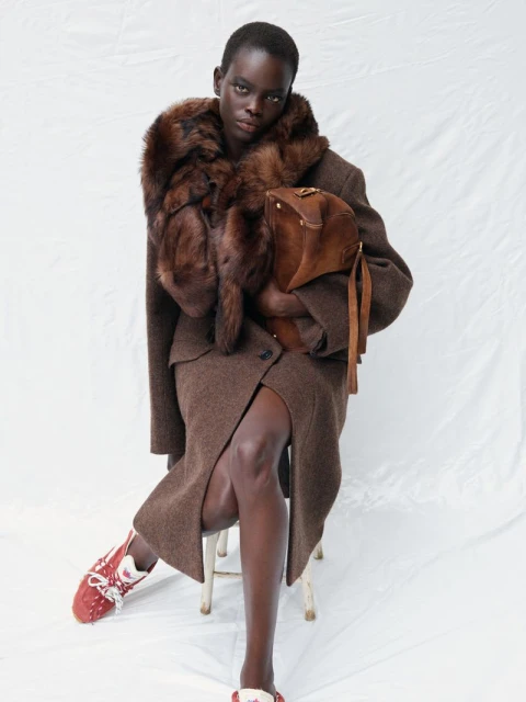 Portrait of a Black fashion model in a brown skirt suit with a fur collar and purse, seated against a white backdrop in a studio.