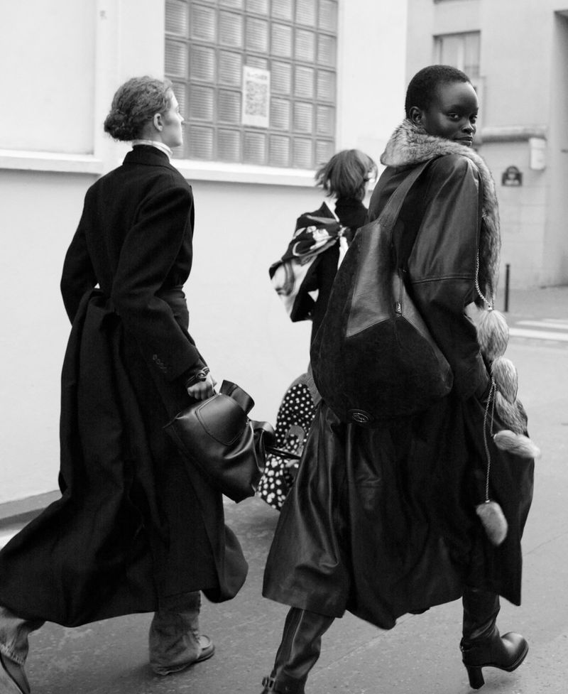 Black and white street style photograph capturing a group of fashionably dressed people walking along a city street, showcasing long coats and boots.