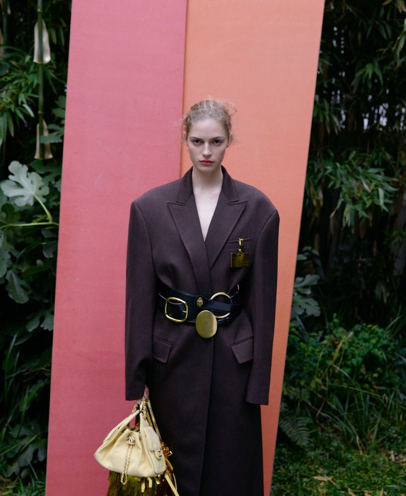 A woman in an oversized brown coat, cinched with a black belt and gold buckle, holds a handbag against a backdrop of pink panels and foliage.