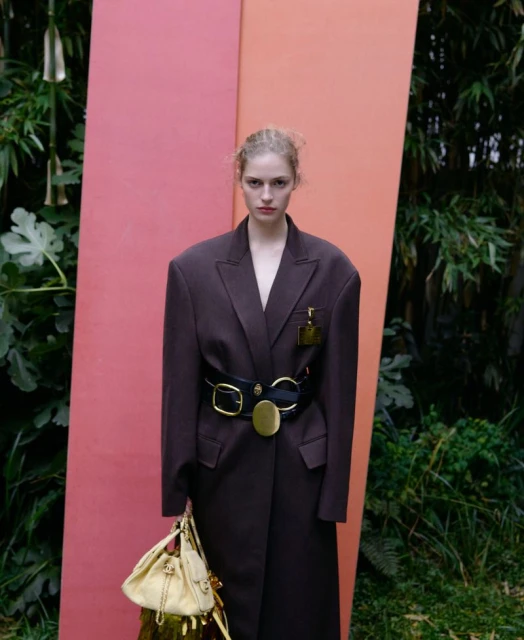 A woman in an oversized brown coat, cinched with a black belt and gold buckle, holds a handbag against a backdrop of pink panels and foliage.