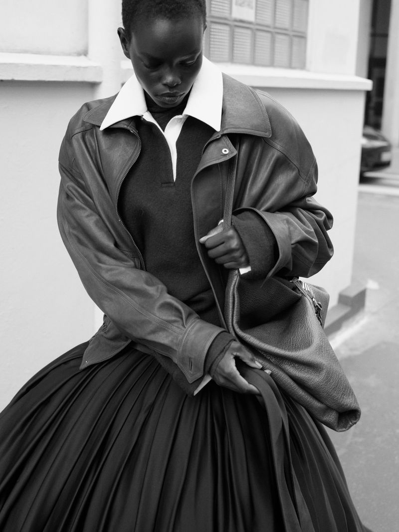 Black and white editorial portrait of a model wearing an oversized leather jacket and pleated skirt, holding a handbag.