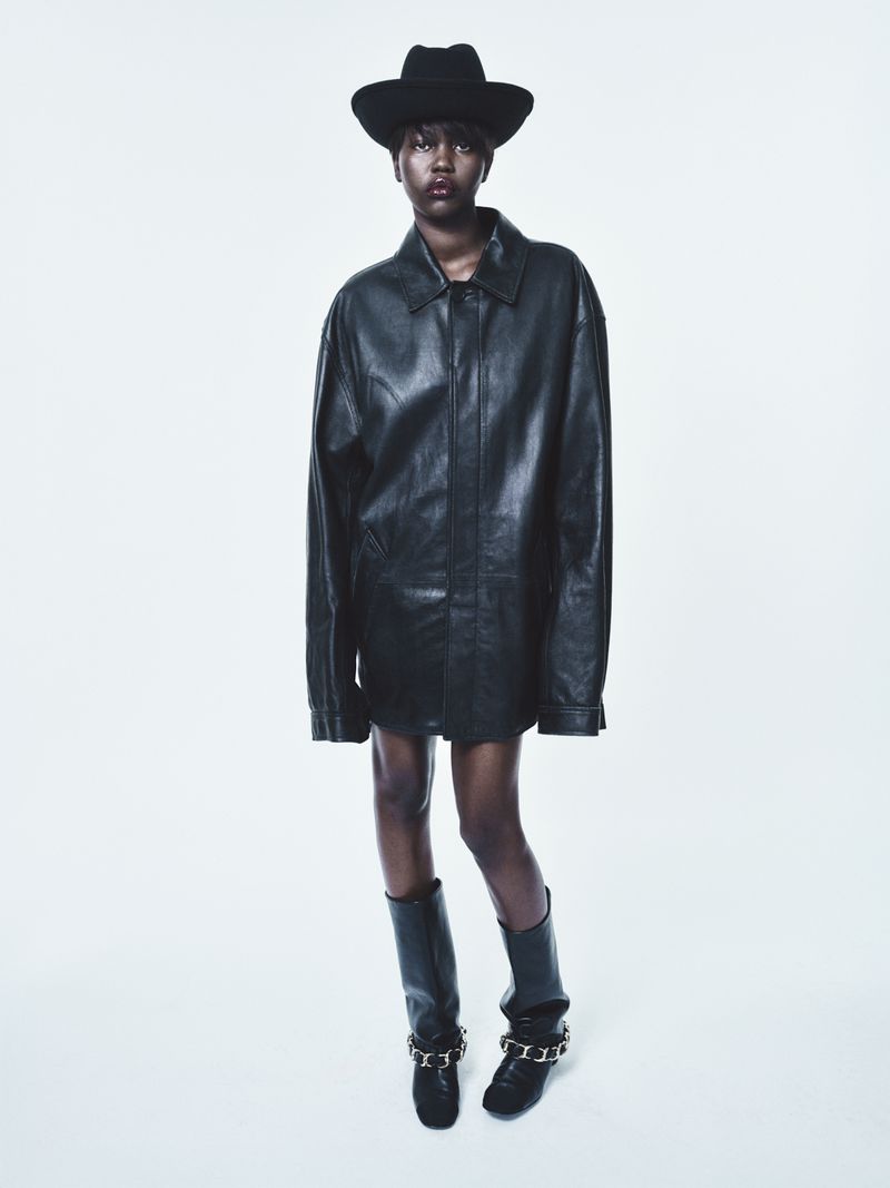 Elegant African-American woman poses in oversized black leather coat, hat, and boots against a bright white backdrop in a fashion setting.