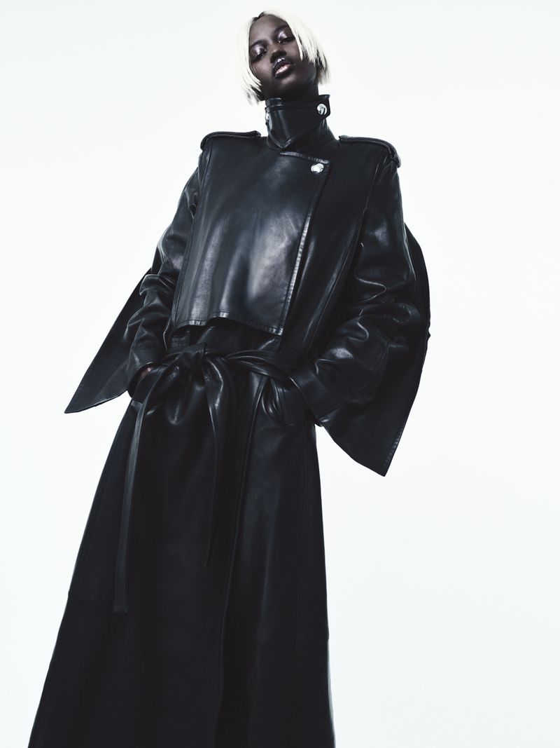 Fashion shot of a Black woman with dyed blonde hair wearing a shiny black leather trench coat against a seamless white backdrop.