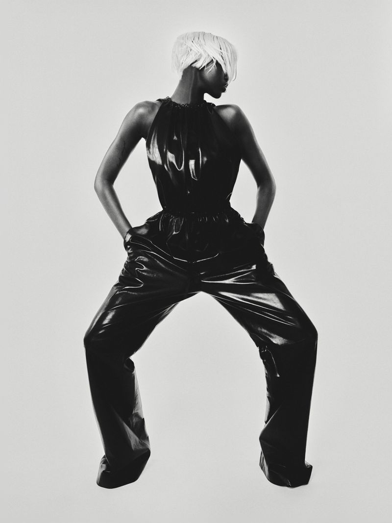 Fashion shot of a Black woman with platinum blonde hair wearing a shiny black top and wide-legged pants against a white background.