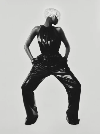 Fashion shot of a Black woman with platinum blonde hair wearing a shiny black top and wide-legged pants against a white background.