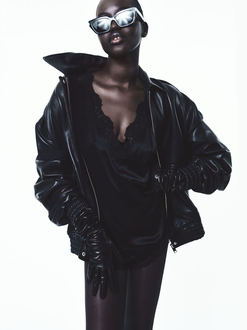Model Adut Akech wearing an oversized black leather jacket, black leather gloves, and reflective sunglasses against a white background.