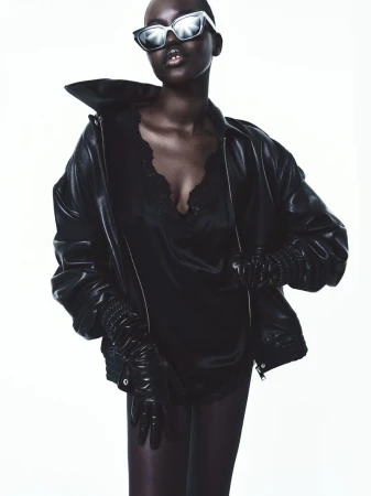 Model Adut Akech wearing an oversized black leather jacket, black leather gloves, and reflective sunglasses against a white background.