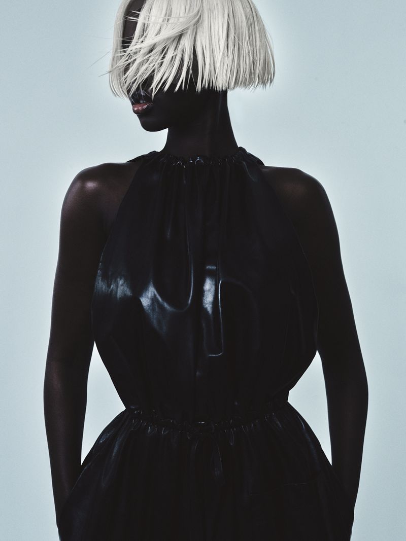 A woman with dark skin and short white hair poses in a black, sleeveless dress against a soft blue background in a studio shot.