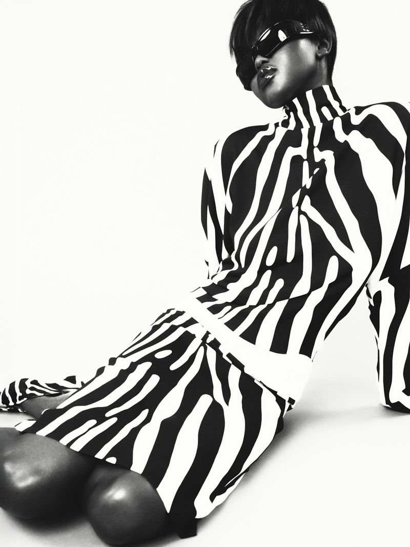 A woman in a black and white zebra-striped dress poses with sunglasses on a white background in a fashion shot with neutral tones.