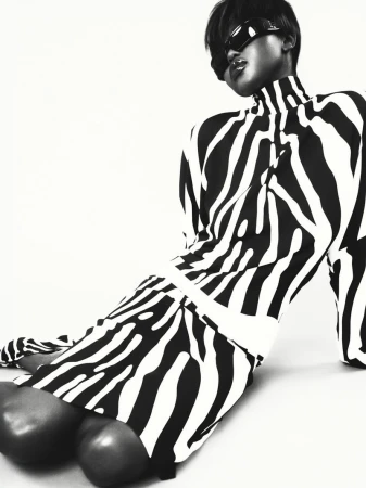 A woman in a black and white zebra-striped dress poses with sunglasses on a white background in a fashion shot with neutral tones.