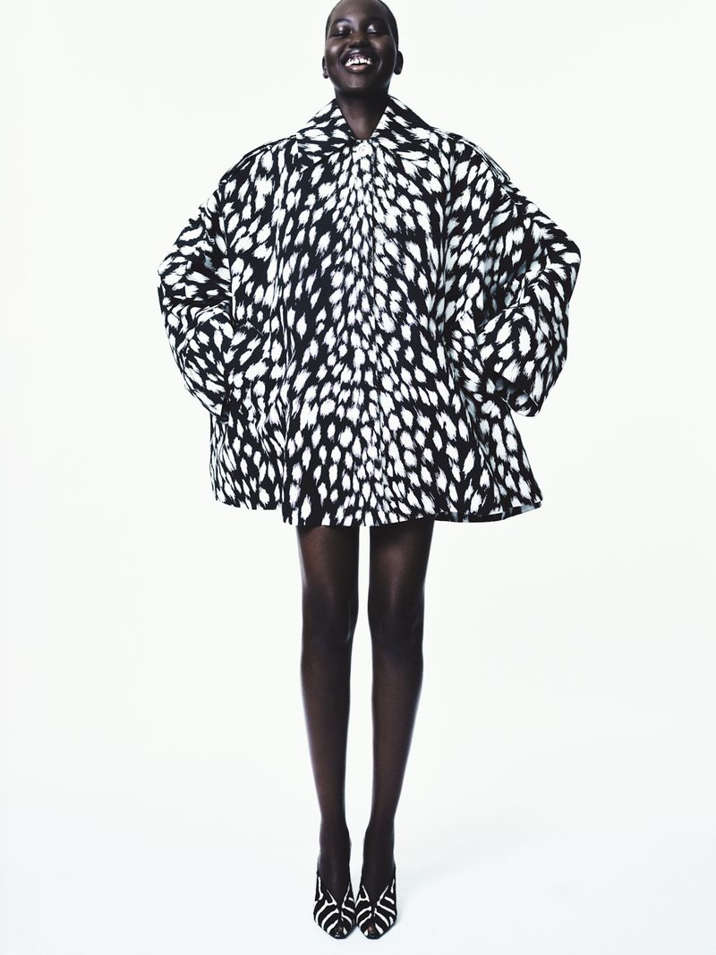 A cheerful black female model poses in a studio setting, showcasing a stylish black and white patterned jacket with matching heels.