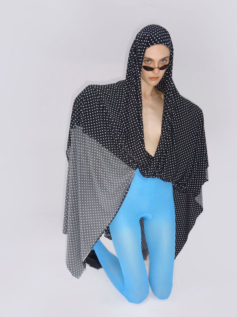 Person kneeling wearing sunglasses, patterned hooded cape, and blue leggings, posing in front of a white background in a studio setting.