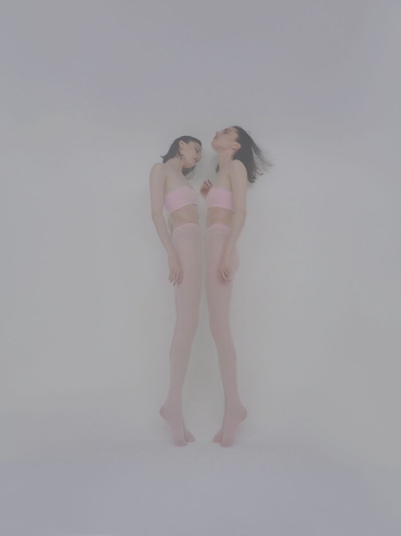 Two women stand in a studio wearing pink tops and tights, with their heads tilted back. The background is a soft white.