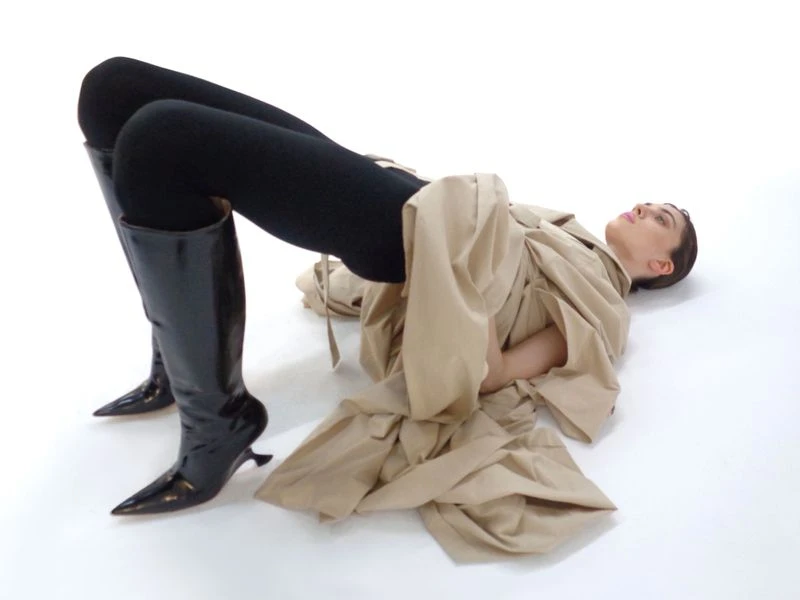 Woman in beige trench coat lying on white background wearing black leggings and high-heeled black patent leather boots.