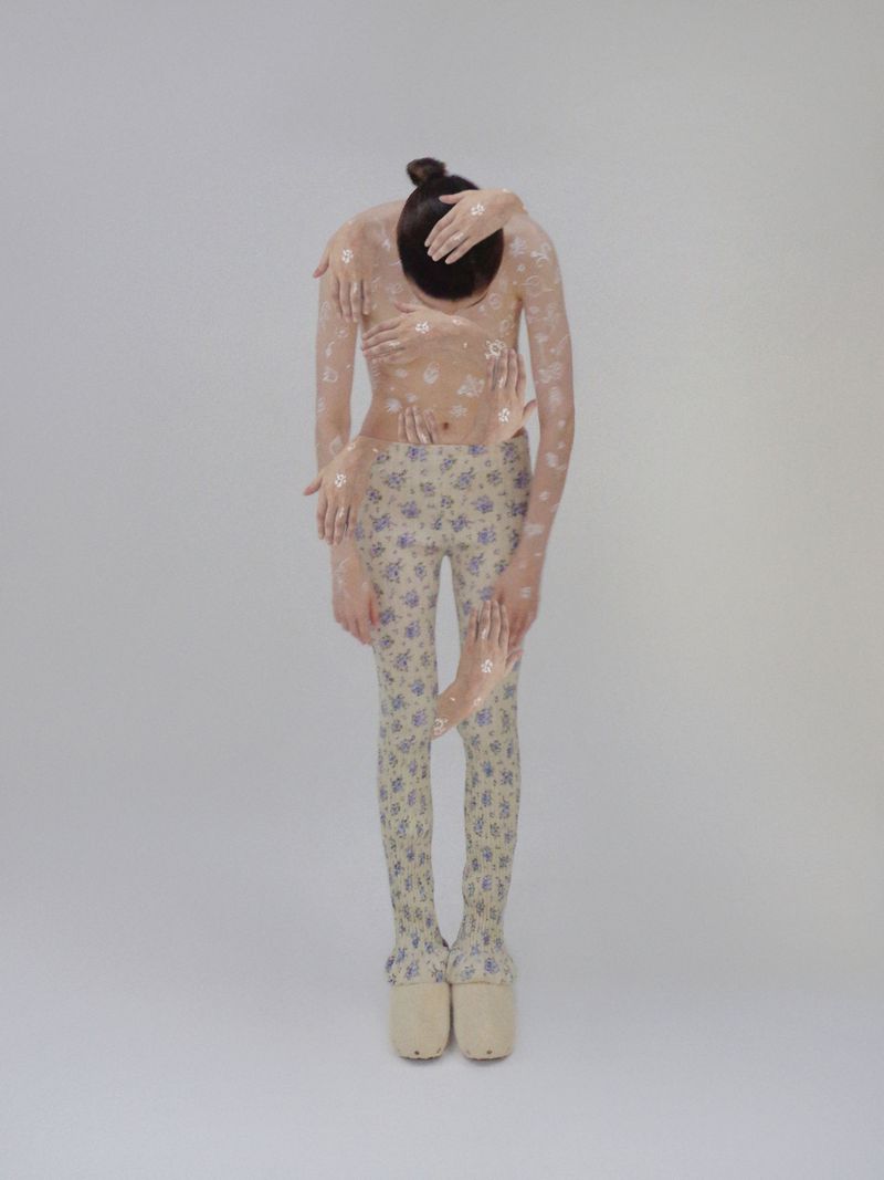 Full-length portrait of an androgynous figure covered in a floral pattern and multiple arms, wearing platform shoes on a white backdrop.