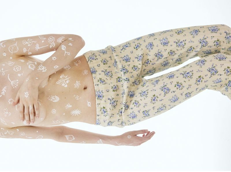 Model lying down wearing floral print trousers with white botanical drawings painted across their torso and arms.