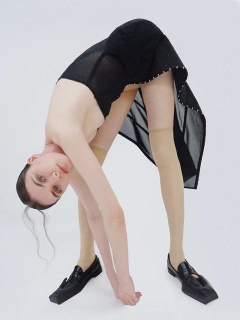 Woman in black dress with semi-sheer details bending over backwards with flat black loafers on a solid white background in studio lighting.