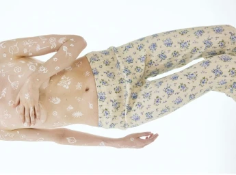 Model lying down wearing floral print trousers with white botanical drawings painted across their torso and arms.