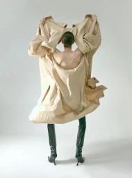Model Milena Litvinovskaya seen from behind holding an oversized beige coat upward while wearing black heeled boots in a grey studio.