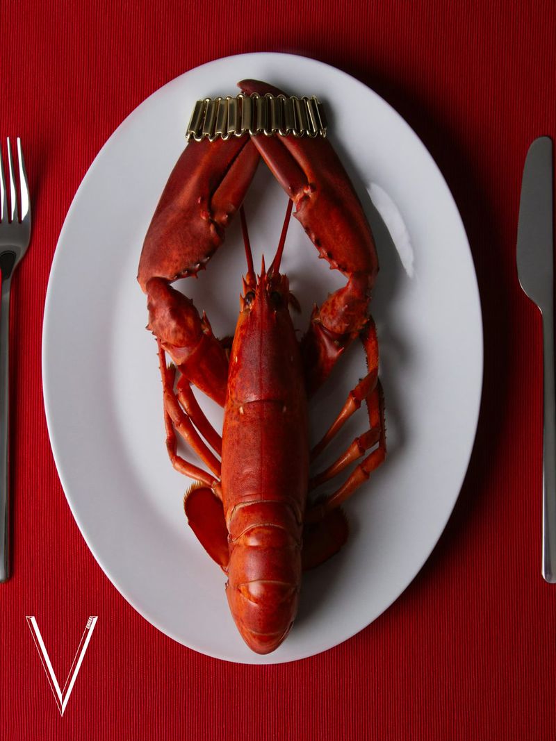 Overhead shot of a cooked red lobster served on a white plate with a gold paper crown, fork, and knife on a red tablecloth.
