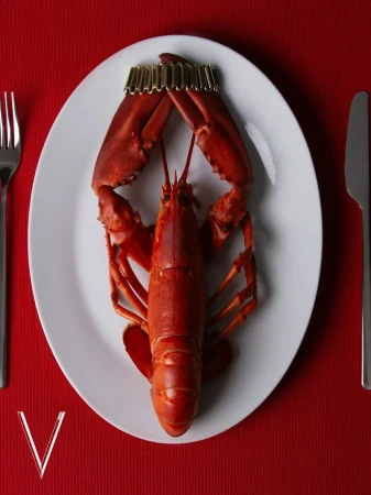 Overhead shot of a cooked red lobster served on a white plate with a gold paper crown, fork, and knife on a red tablecloth.