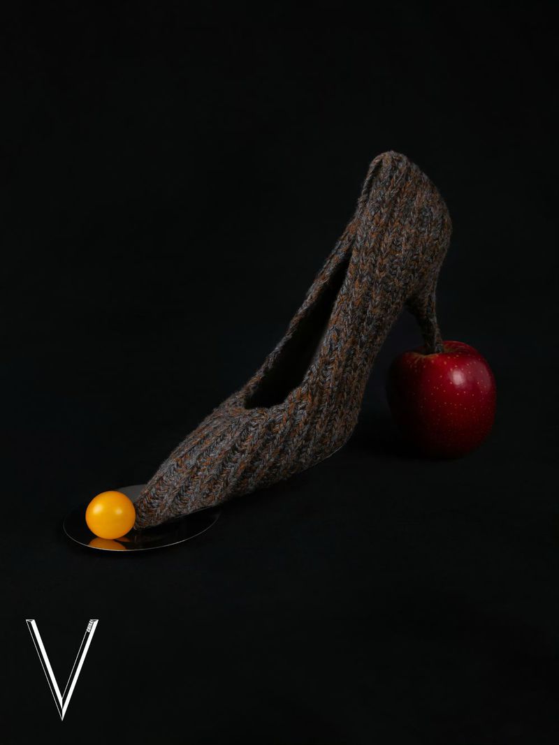 High heel shoe made of textile material next to a red apple and orange ball on a black background.