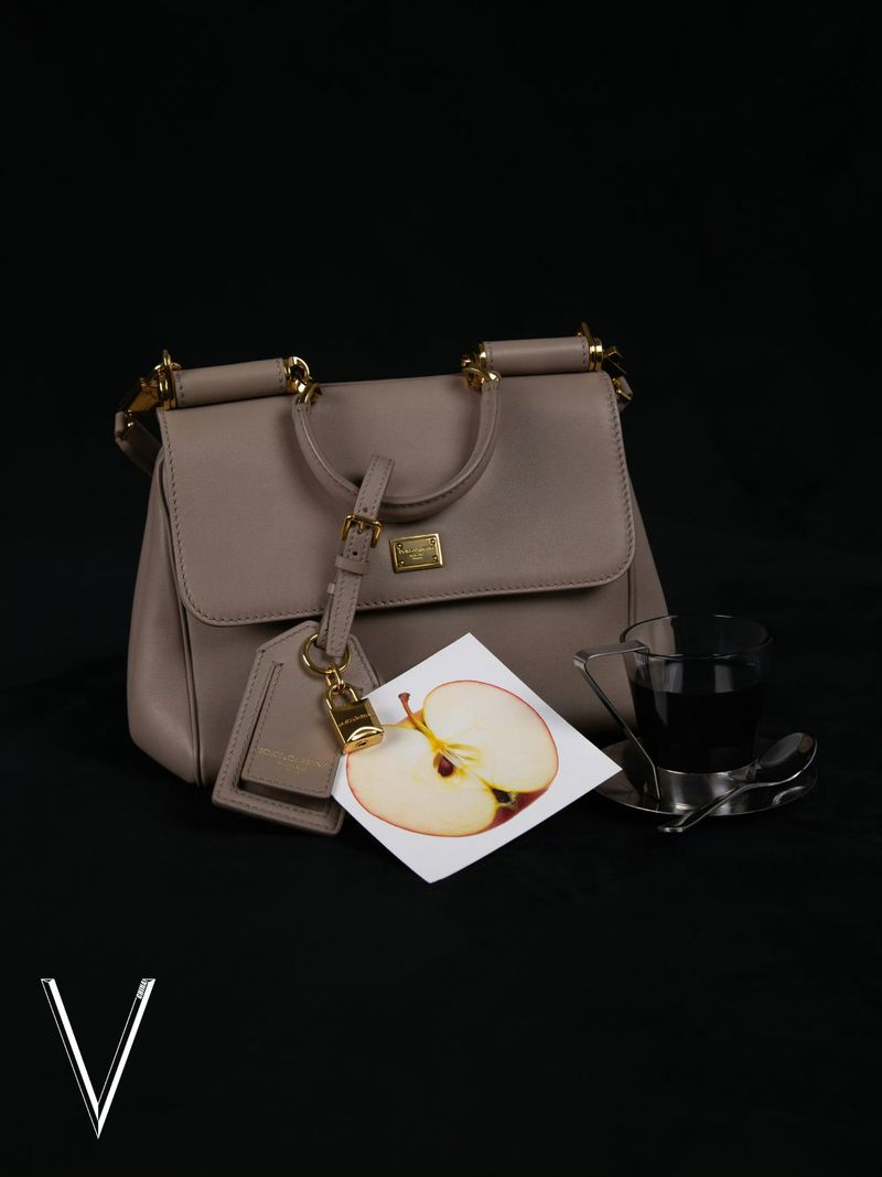 Elegant taupe handbag with gold accents on a black background, complemented by a decorative apple card and a sleek glass.