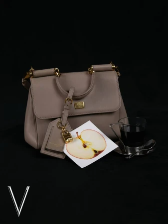 Elegant taupe handbag with gold accents on a black background, complemented by a decorative apple card and a sleek glass.