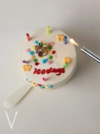 Close-up of a decorated white cake with candles being lit with a lighter. The cake has red text spelling '1000days'.