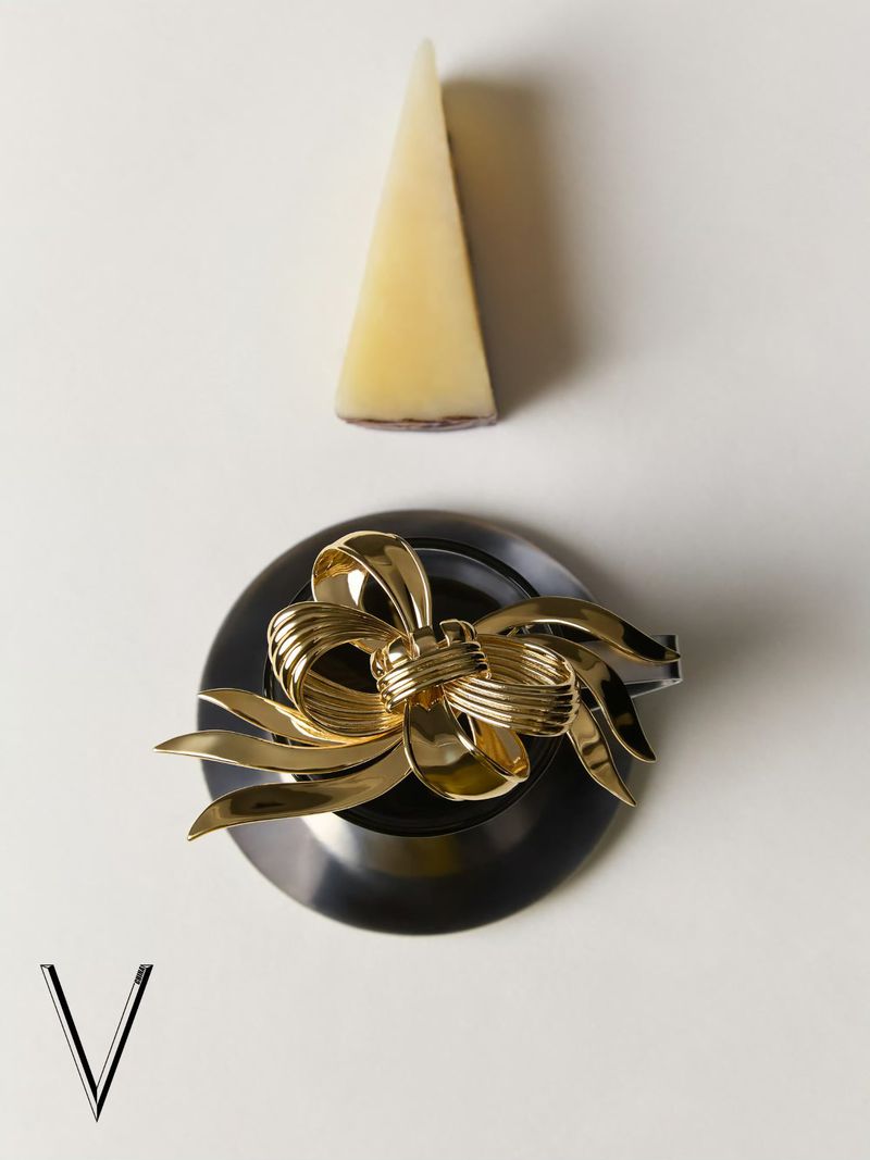 Overhead shot of a gold bow brooch on a black plate, next to a triangular slice of cheese, set against a white surface.