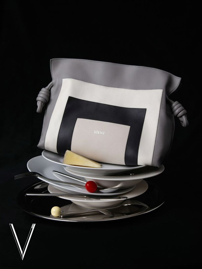 Fashion still life of a gray Loewe handbag on top of a stack of white plates with silverware against a black background in studio lighting.