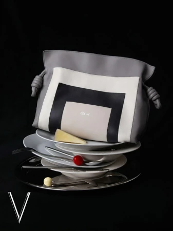 Fashion still life of a gray Loewe handbag on top of a stack of white plates with silverware against a black background in studio lighting.