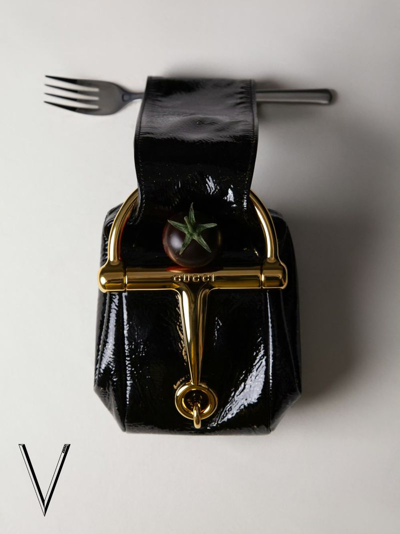 Patent-leather black Gucci handbag held up by a silver fork against a plain background with a dark tomato inside.