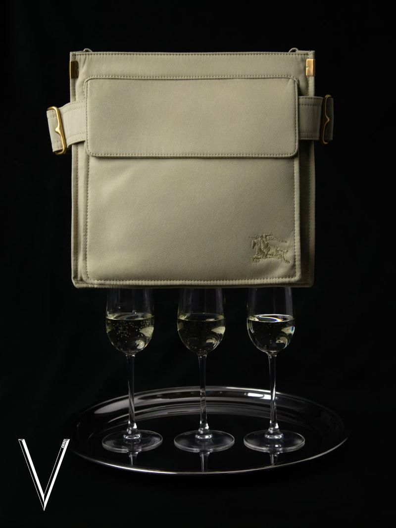 Beige satchel-style purse floating above three champagne flutes on a silver tray, set against a dark background in a studio shot.