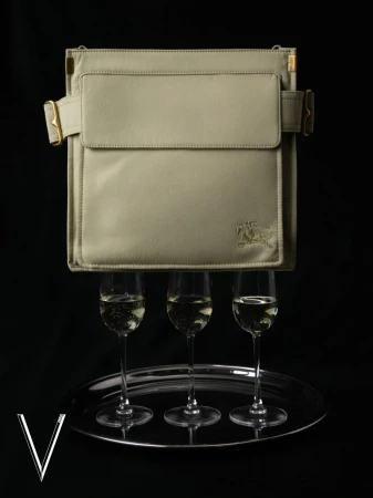 Beige satchel-style purse floating above three champagne flutes on a silver tray, set against a dark background in a studio shot.