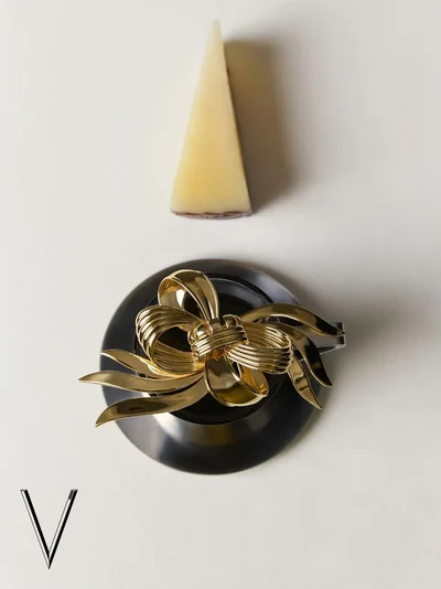 Gold bow brooch resting on a dark object with a triangular cheese wedge above on white surface