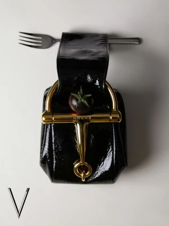 Black patent leather Gucci horsebit bag styled with a fork and dark cherry tomato against an off-white background.