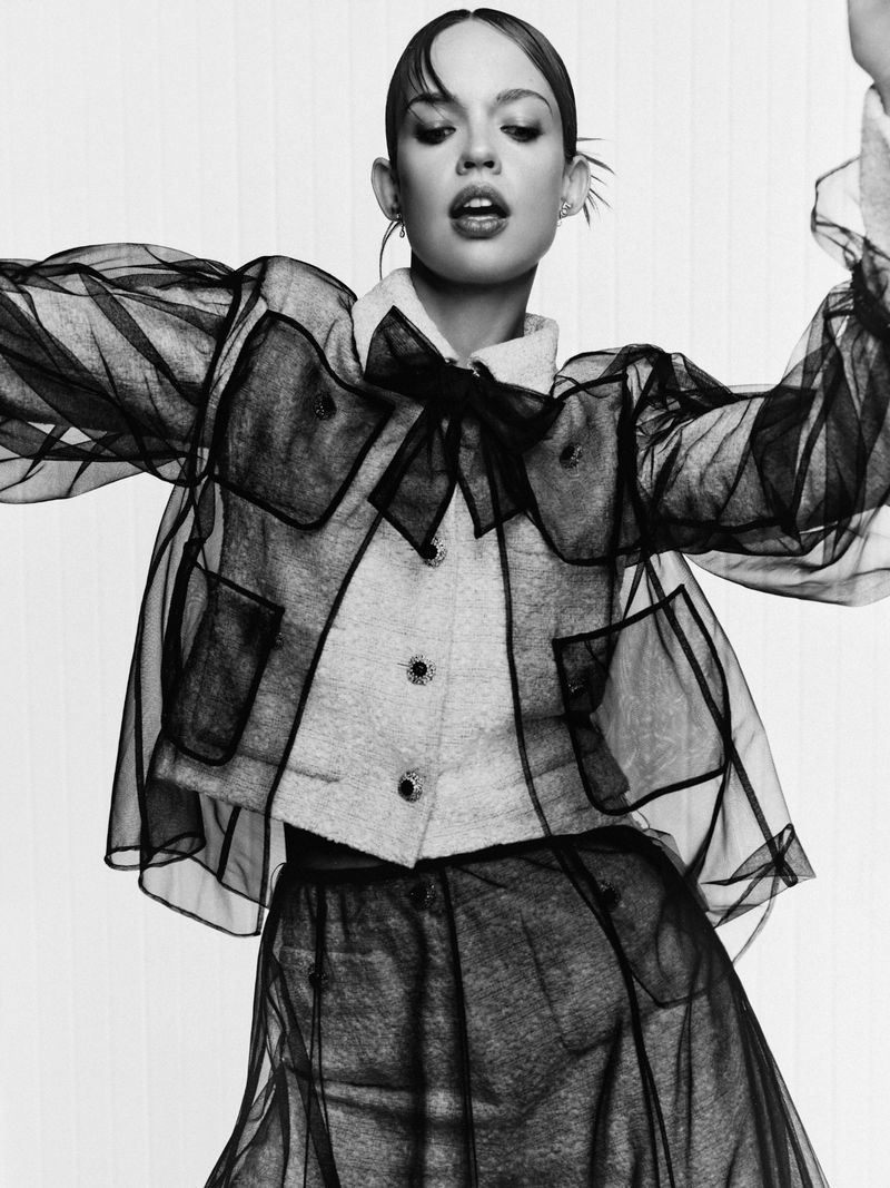A young woman poses in a grey collared jacket with black trim, along with a sheer black jacket, wrist-length sleeves, and matching skirt.