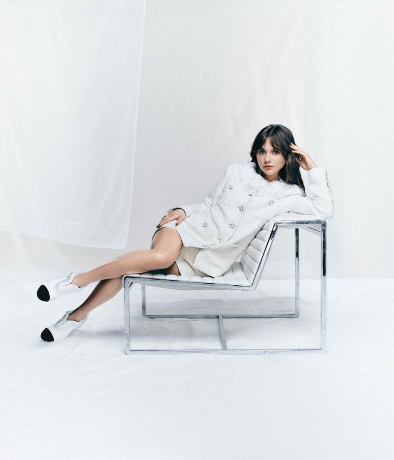 A woman with brown hair lounges gracefully in a modern metal chair, wearing a white jacket and shoes, against a bright white backdrop.