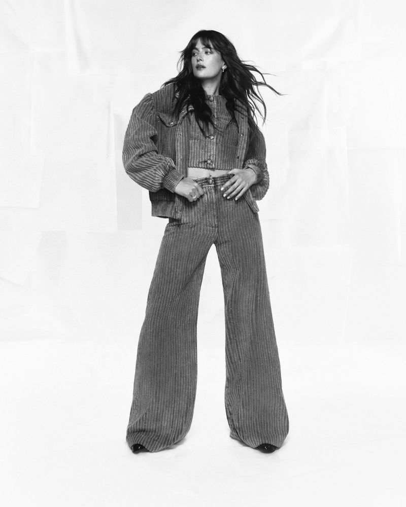 A young woman with flowing brown hair poses in a three-piece, striped, corduroy outfit against a stark white background, showcasing fashion and style.