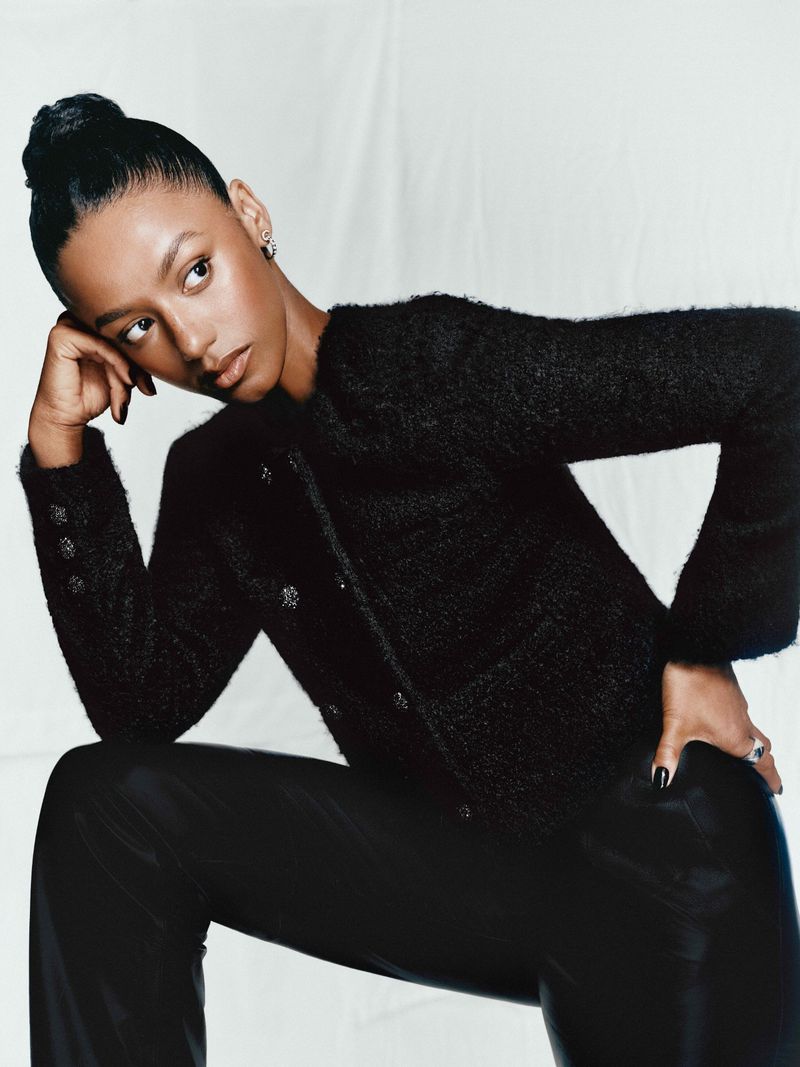 A woman poses in a dark, textured cardigan and leather-look pants against a white background, striking a stylish pose with her hand near her face.